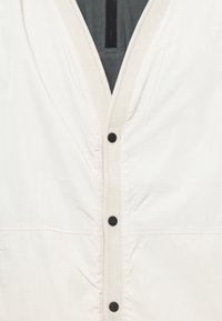 Lightweight cream jacket with a deep V-neck, fabric snap buttons, and subtle texture, featuring a contrasting dark lining.