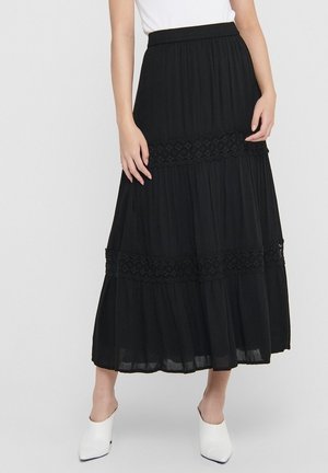 Zwarte maxirok met lagen en kanten rand, gedragen met een wit topje en witte mules met puntige neus, staand tegen een effen achtergrond.