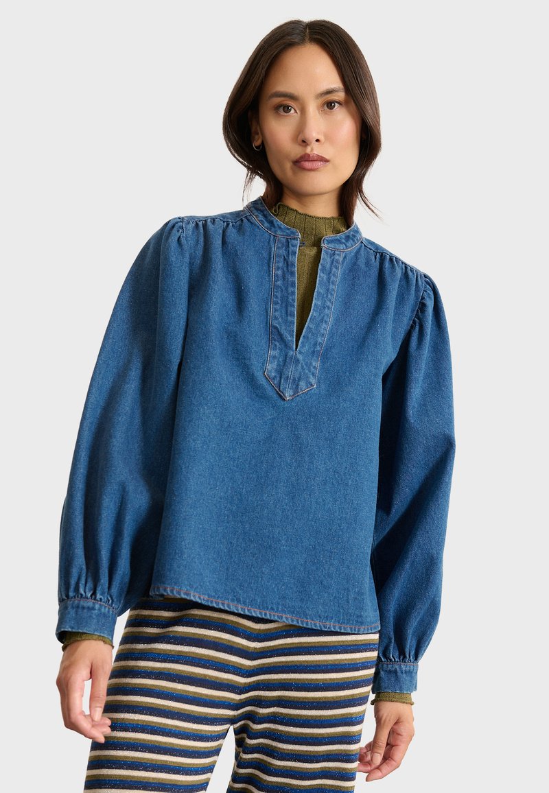 Denimbluse in Blau mit lockerer Passform, puffärmel und einer gekerbten Halslinie. Kombiniert mit gestreiften, strukturierten Hosen in verschiedenen Farben.