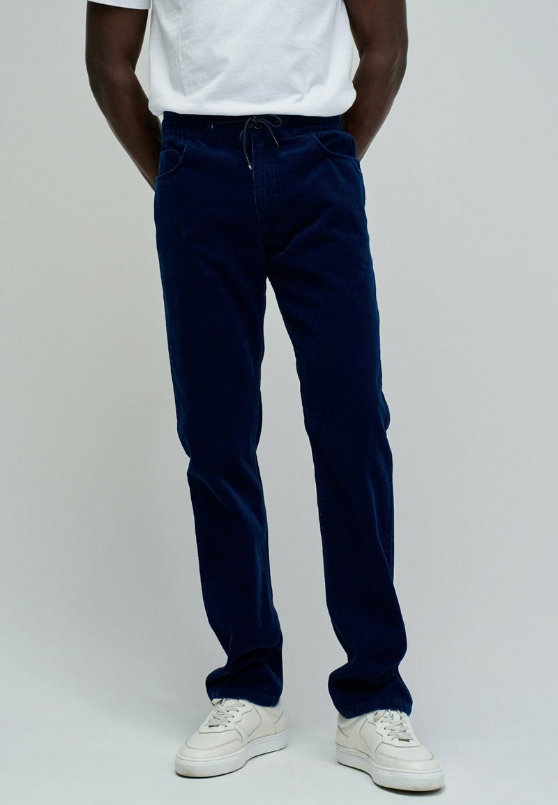 Pantalons en velours côtelé bleu marine avec une taille à cordon, deux poches avant et une coupe droite, associés à des baskets blanches et un t-shirt blanc uni.