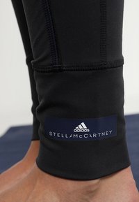 Svarta leggings i stretchigt material, med en marinblå logotag som har "adidas" och "STELLA McCARTNEY" vid vristen.