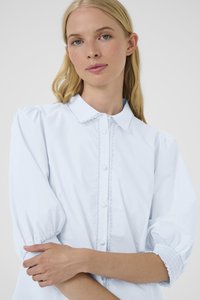 Donna bionda che indossa una blusa azzurro chiaro con maniche a sbuffo, bordi ondulati lungo i bottoni e il colletto, braccia incrociate in vita.