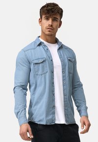 Joven con cabello rizado corto que lleva una camisa de mezclilla azul clara sobre una camiseta blanca, de pie contra un fondo claro y liso.