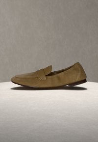 Καφέ σουέντ μοκασίνι με χαμηλό τακούνι, στρογγυλή μύτη και λεπτομέρεια με λουρί, παρουσιασμένο σε ουδέτερη επιφάνεια με απλό φόντο.