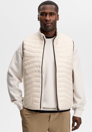 Man die een lichtbeige gewatteerd vest draagt over een crèmekleurige trui, met kaki broek, staand tegen een effen witte achtergrond.