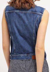 Bez rękawów kurtka dżinsowa w jasnoniebieskim odcieniu, klasycznym kroju i zamszową metką na tylnej krawędzi. Tkanina o fakturze z widocznymi szwami.