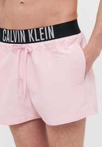 Helle rosa Badehosen mit elastischem schwarzen Bund, auf dem "CALVIN KLEIN" in weißer Schrift steht. Mit Kordelzug und seitlichen Taschen.