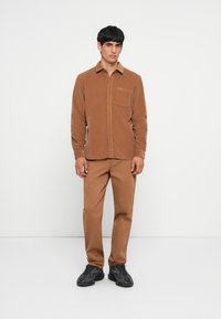 Brown corduroy shirt with buttons, chest pocket, paired with matching brown pants and black sneakers, standing on a white background.