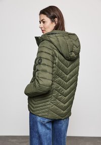 Doudoune verte olive avec capuche, arborant un motif en chevrons et une accent logo sur la manche. Portée sur un denim, affichée de dos.
