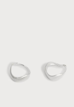 Paire de boucles d’oreilles créoles lisses couleur argent avec une forme triangulaire légèrement irrégulière sur un fond clair uni.