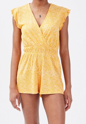 Mujer con mono corto amarillo de manga corta con cintura elástica y delicadas mangas con volantes, combinado con collares de plata en capas.