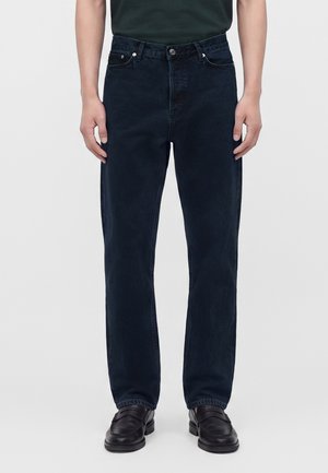 Homme portant un jean bleu foncé droit et des chaussures en cuir noires, debout devant un fond uni.