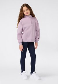 Fille jeune avec de longs cheveux bouclés portant un sweat à capuche lilas zippé, des leggings navy et des baskets blanches, se tenant devant un fond uni.