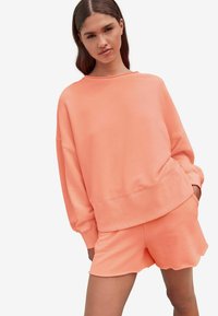 Koralfarvet sweatshirt med brede manchetter og en afslappet pasform, parret med shorts med bølget kant i samme farve. Glat tekstur, afslappet design.