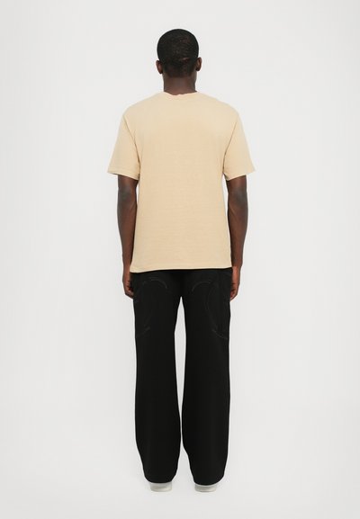 T-shirt en coton beige avec une coupe décontractée, associé à un pantalon noir ample avec des coutures ton sur ton et un design minimaliste.