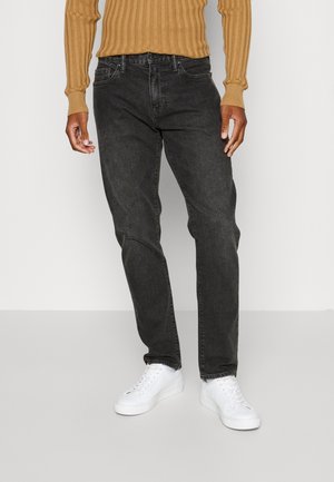 Mężczyzna w czarnych dżinsach, białych trampkach i brązowym prążkowanym swetrze z długim rękawem, stojący na tle jednolitego jasnego tła.