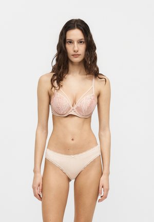 Jeune femme aux longs cheveux bruns, debout face à l'avant, portant un soutien-gorge en dentelle rose clair et une culotte taille haute assortie sur un fond uni.