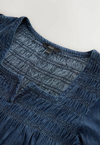 Denim majica s V-izrezom i nabranim detaljem. Ima teksturiranu, mekanu plavu tkaninu s obrubljenim rubom i vidljivom oznakom brenda.