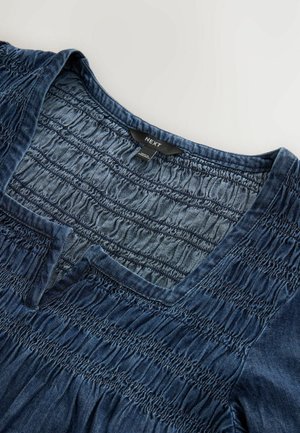 Denimowa bluzka z dekoltem w kształcie litery V i zaszewkami. Posiada teksturowany, miękki niebieski materiał z wykończonym dołem oraz widoczną metką marki.