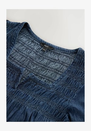 Denimowa bluzka z dekoltem w kształcie litery V i zaszewkami. Posiada teksturowany, miękki niebieski materiał z wykończonym dołem oraz widoczną metką marki.