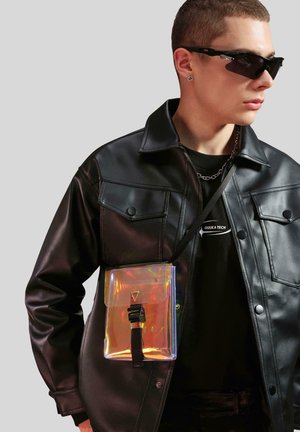 Transparent holografisk taske med flaplukning, en sort rem, parret med en sort læderjakke og en sort grafisk T-shirt.