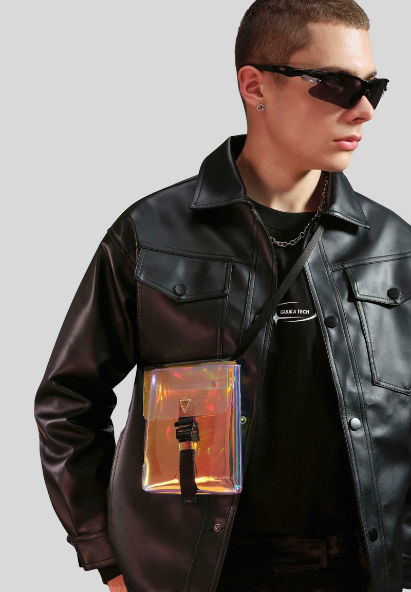Transparent holografisk väska med lockstängning, ett svart rem, matchad med en svart skinnjacka och en svart grafisk T-shirt.