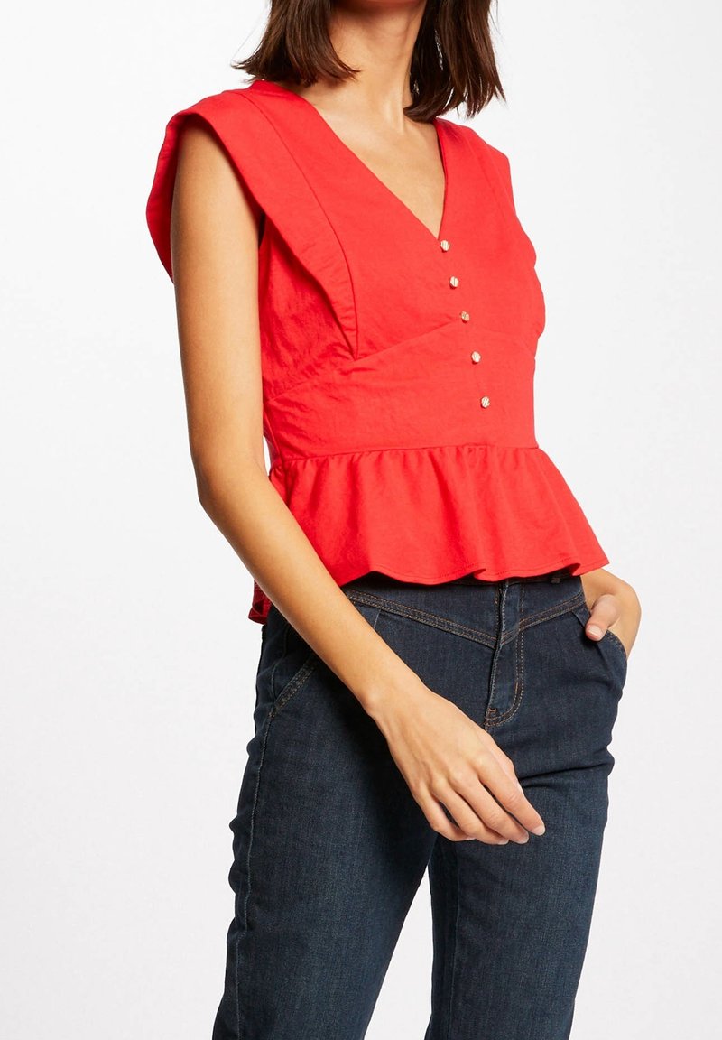 Femme portant un haut peplum rouge sans manches avec des boutons décoratifs et un jean bleu foncé, une main dans la poche, sur fond blanc.