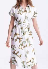 Una donna che indossa un vestito midi bianco con motivi floreali verdi e marroni, maniche corte, colletto e una cintura coordinata in vita.