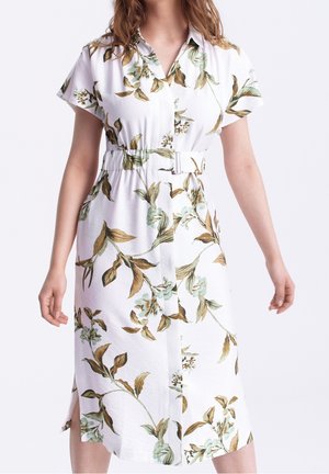 Femme portant une robe midi blanche avec des motifs floraux verts et bruns, à manches courtes, col et ceinture assortie à la taille.