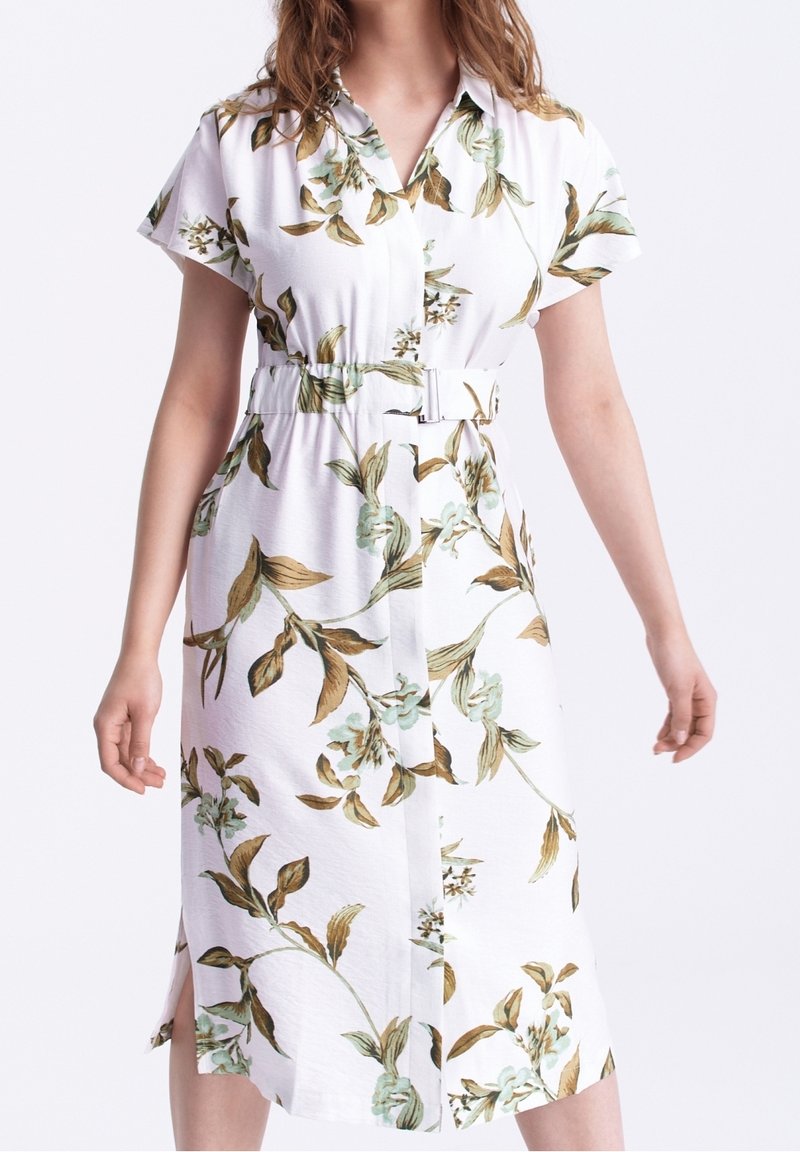 Una donna che indossa un vestito midi bianco con motivi floreali verdi e marroni, maniche corte, colletto e una cintura coordinata in vita.