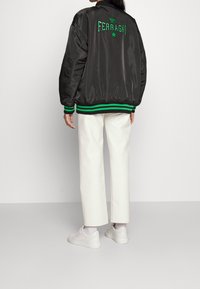 Svart satin bomberjacka med grön broderad text och stjärna på ryggen, med kontrasterande grönrandiga manchetter och nederdel. Bärs med vita byxor.