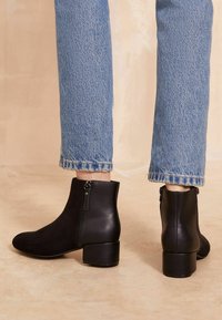 Persona vestida con jeans azules y botas negras de tobillo con tacones cuadrados y cremalleras laterales, de pie sobre un suelo beige.
