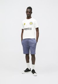 Bílé tričko s potiskem a žlutými akcenty v kombinaci s fialovými šortkami. Černé tenisky a bílé ponožky doplňují ležérní outfit.