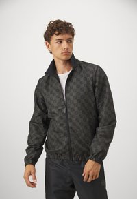 Giacca con zip, a colletto alto, nera e verde scuro, con motivo a fantasia. Maniche con polsini elasticizzati e cintura. Texture liscia con un design geometrico.