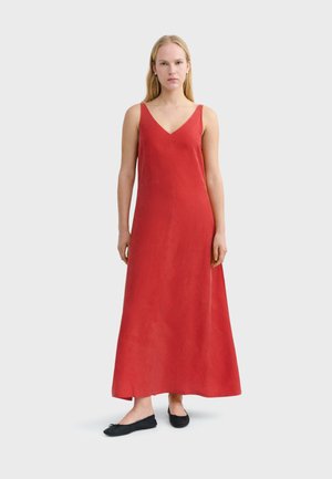 Femme debout portant une robe maxi rouge sans manches avec un décolleté en V et des chaussures plates noires sur un fond clair uni.