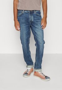 Jeans de denim azul con corte recto, que presentan bolsillos clásicos cosidos, un lavado desgastado y un dobladillo enrollado. Combinados con zapatillas de colores múltiples.