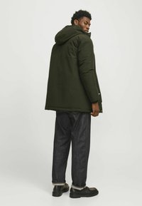 Tume roheline soojustatud mantli koos kapuutsiga, sirge lõikega, küljetaskute ja lukuga, koos tume denimi pükste ja mustade kingadega.