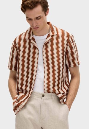 Mann in kurzärmeligem braun-weiß gemustertem Hemd über weißem T-Shirt, trägt beige Hose, Hände in den Taschen, schaut nach unten vor einfarbigem Hintergrund.