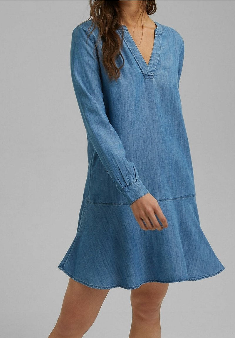 Femme portant une robe en denim bleue décontractée, ample, à manches longues et col en V, debout devant un fond gris uni.