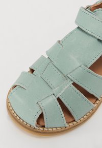 Mintgrønn lærsandal med flettet design, åpen tå og justerbar stropp. Merkbare sømdetaljer på sålen og overdelen.
