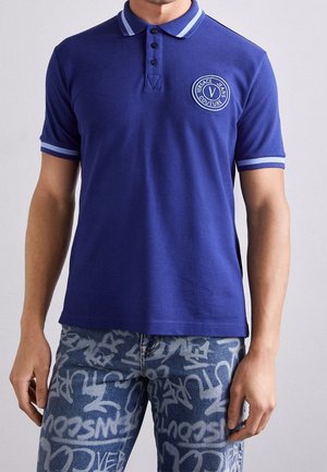 Man wearing blue polo shirt with light blue trim and Versace Jeans Couture logo patch, paired with blue jeans featuring white graffiti text.