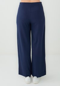Navy blauwe wijde broek van soepel materiaal. Heeft een hoge taille en een schone afwerking langs de zoom. G Draagt met witte sneakers.