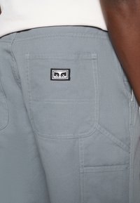 Nahaufnahme einer grauen Hosentasche mit einem schwarz-weißen rechteckigen Logo-Patch, getragen von einer Person in weißem Hemd.