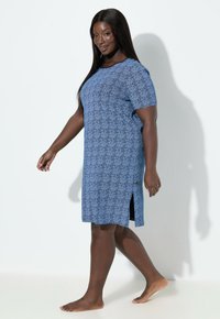 Modré šaty s vzorem, krátkými rukávy, kulatým výstřihem, bočními rozparky a hladkou texturou látky. Modelka je bosá a kráčí po světlém povrchu.