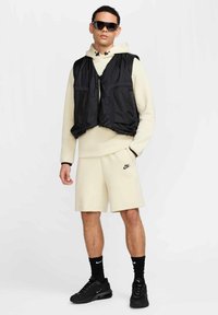 Sort polstret vest over en lys beige hoodie, parret med beige shorts og sorte sneakers. Tilbehør inkluderer mørke solbriller og sorte sokker.