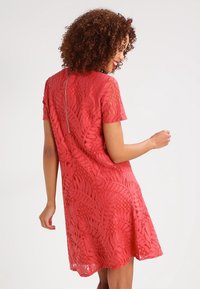 Kobieta z kręconymi włosami w koralowej koronkowej sukience na krótki rękaw z zamkiem błyskawicznym z tyłu, lekko obrócona w bok z uśmiechem.