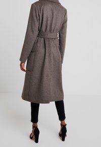 Brauner Wollmischungs-Mantel mit tailliertem Gürtel, großen Taschen und Reverskragen, kombiniert mit schwarzen, figurbetonten Hosen und Absatzschuhen.