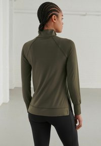 Top de culoare verde închis, cu mâneci lungi și guler fals, cu mâneci raglan și deschideri pe laterale. Textură netedă și design ajustat, asortat cu colanți negri.