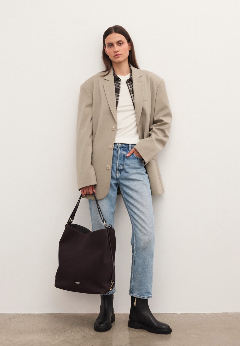 Sieviete stāv ar roku kabatā, ģērbusies pārāk lielā pelēkā mētelī, baltā topā, gaiši zilās džinsās, melnās zābakos, tur liela melna rokassoma.