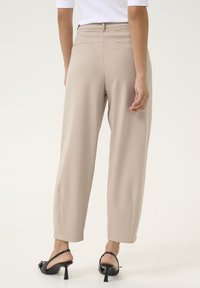 Pantalons beiges ajustés à taille haute, avec poches latérales et jambe fuselée, associés à des sandales à talons noires sur un fond clair.